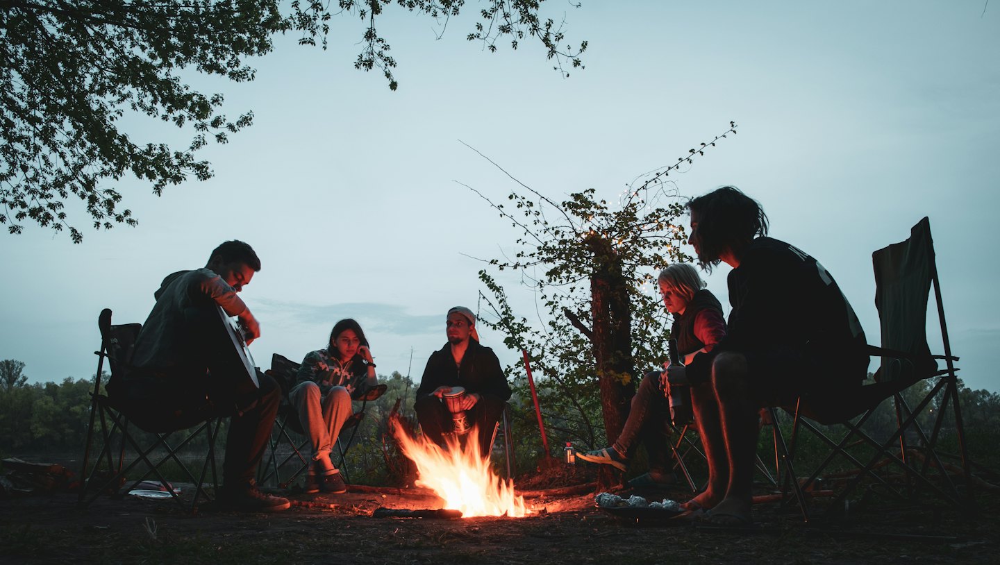 Friends play music and relax around a glowing campfire outdoors.