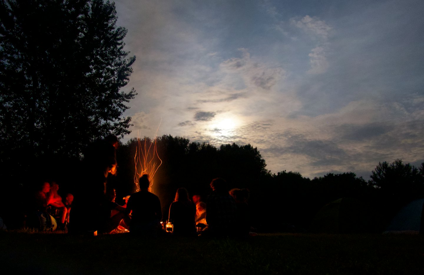 People silhouetted around a glowing campfire at dusk with sparks rising.