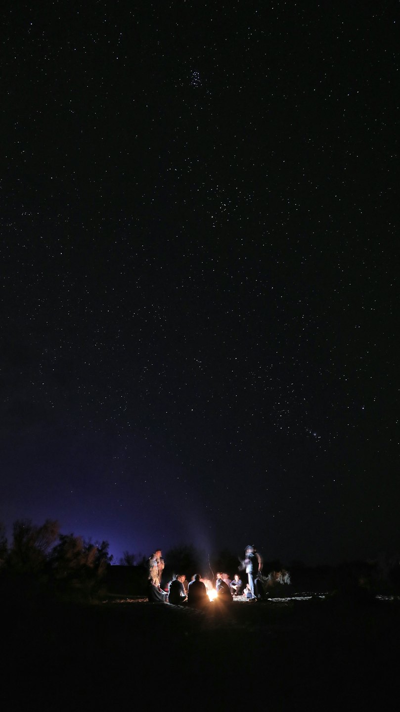 Silhouetted people gather around a warm campfire at night.